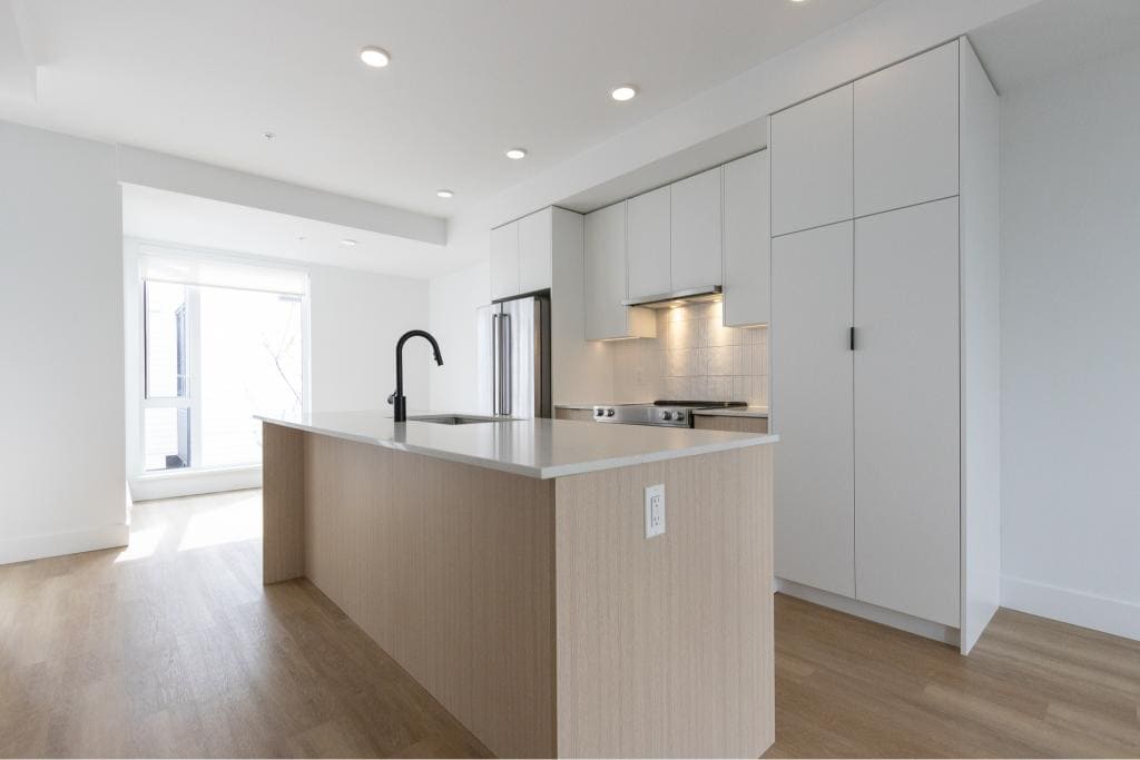 Modern minimalist kitchen in a new condo, featuring white cabinetry, a spacious island with a built-in sink, stainless steel appliances, and natural light. Perfect for urban living in Victoria, BC.