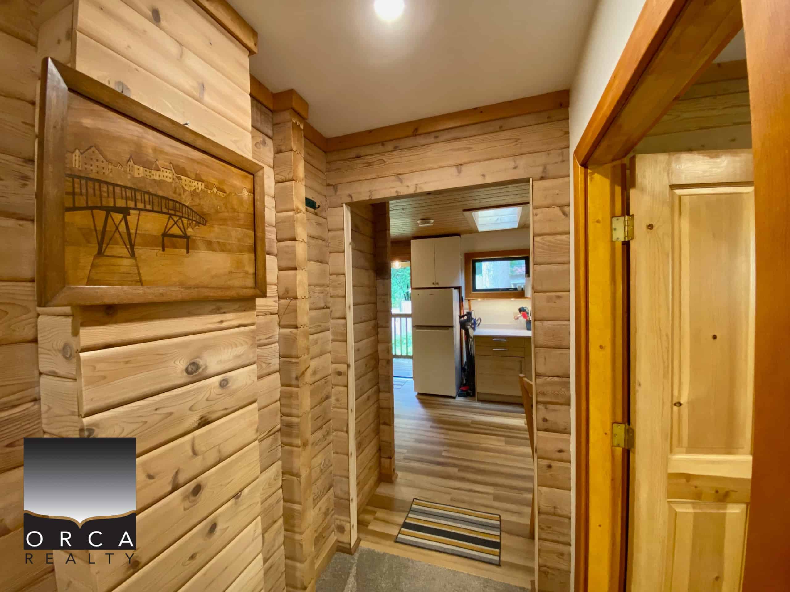 Cozy wooden interior of a home with knotty pine paneling, showcasing warm natural tones, accented by a lake or mountain scene art piece, leading to a kitchen with modern appliances.