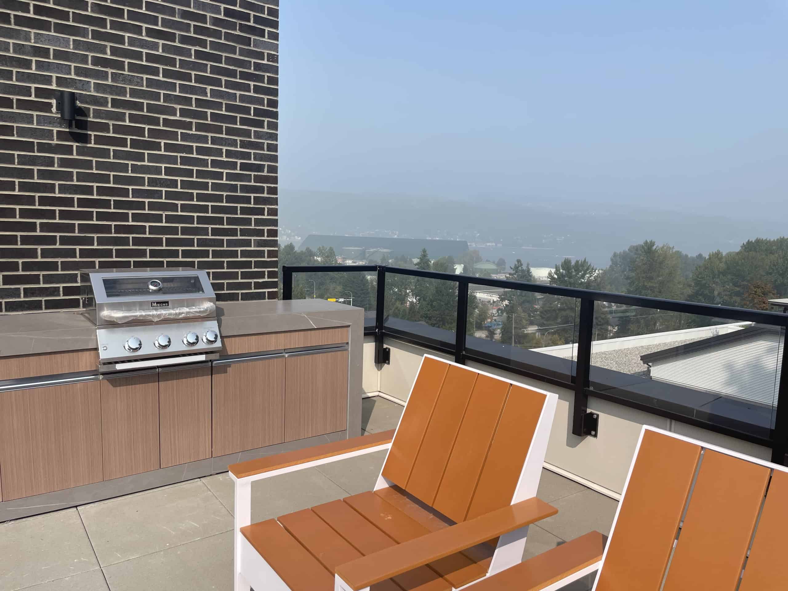 Stunning rooftop balcony with built-in BBQ, modern lounge chairs, and panoramic views of surrounding cityscape and distant mountains, perfect for outdoor entertaining and relaxation in Vancouver.