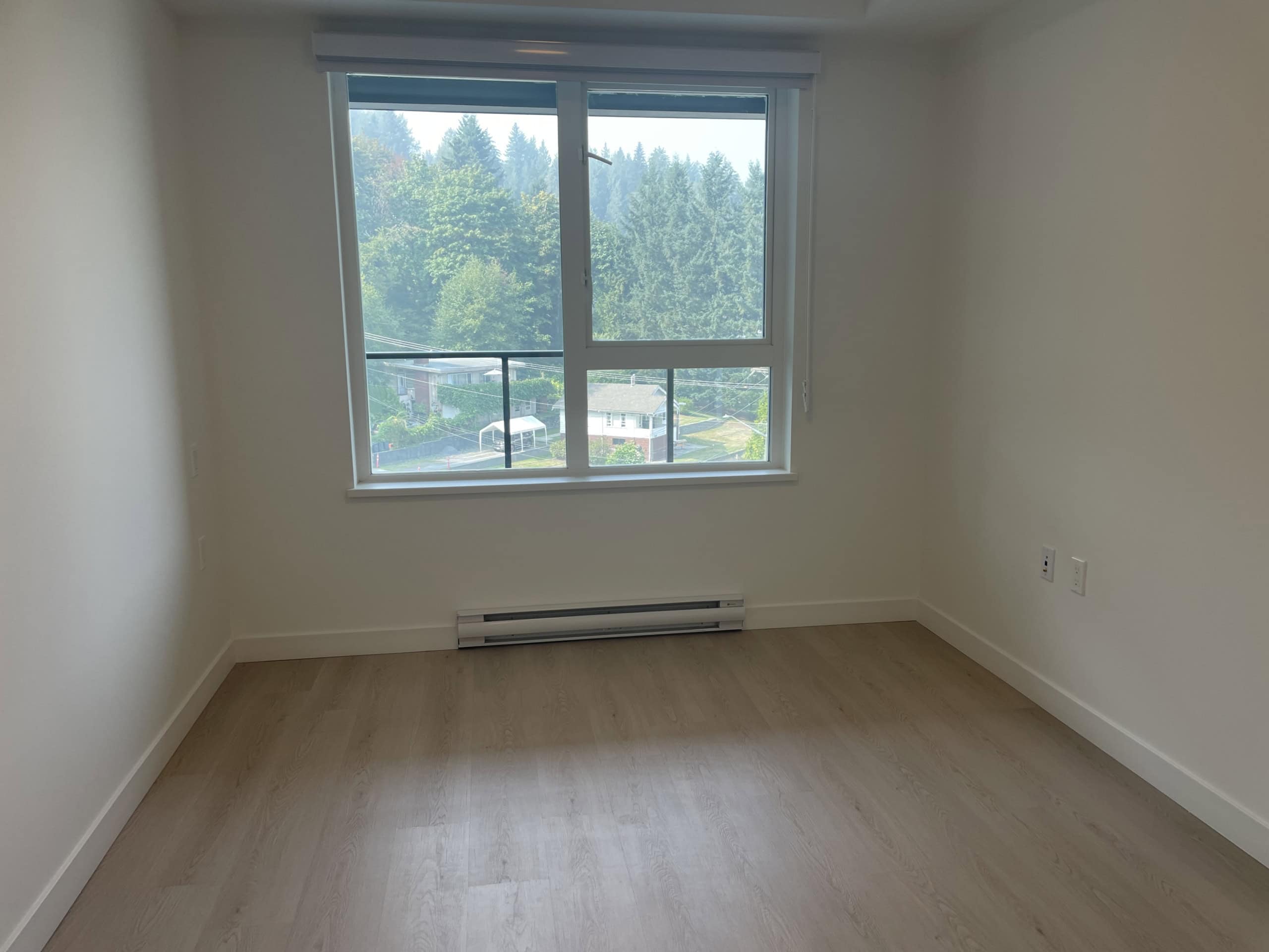 Bright vacant bedroom with large window showcasing lush green trees, modern white walls, and light wood flooring, ideal for in-city living or mountain views at Orca Realty Inc.