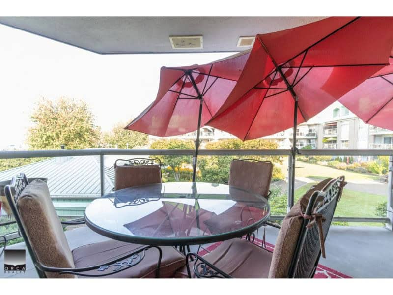 Enjoy outdoor living with a spacious balcony featuring a glass patio table, red umbrellas, and comfortable chairs in a vibrant neighbourhood—perfect for relaxing and entertaining.