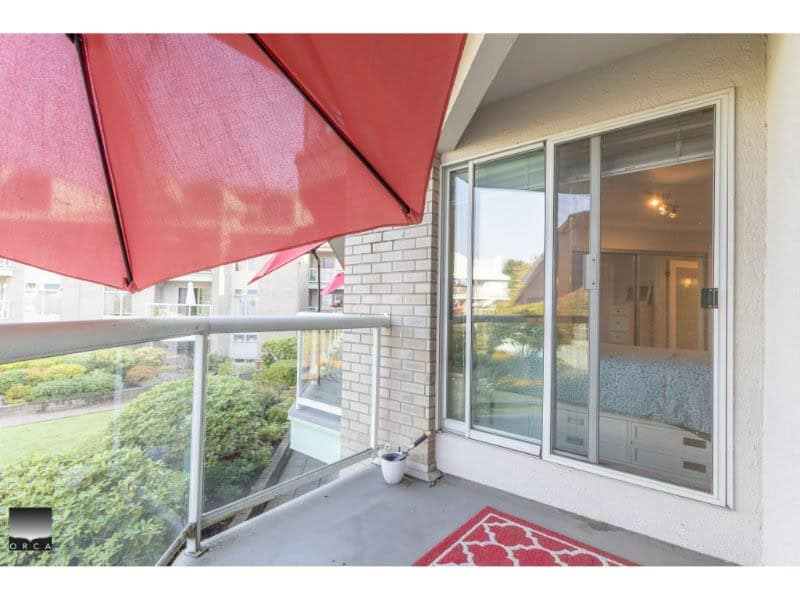 Balcony with red umbrella overlooking lush greenery, sliding glass doors leading to a bright, modern apartment in Vancouver. Perfect for relaxing and enjoying scenic views.