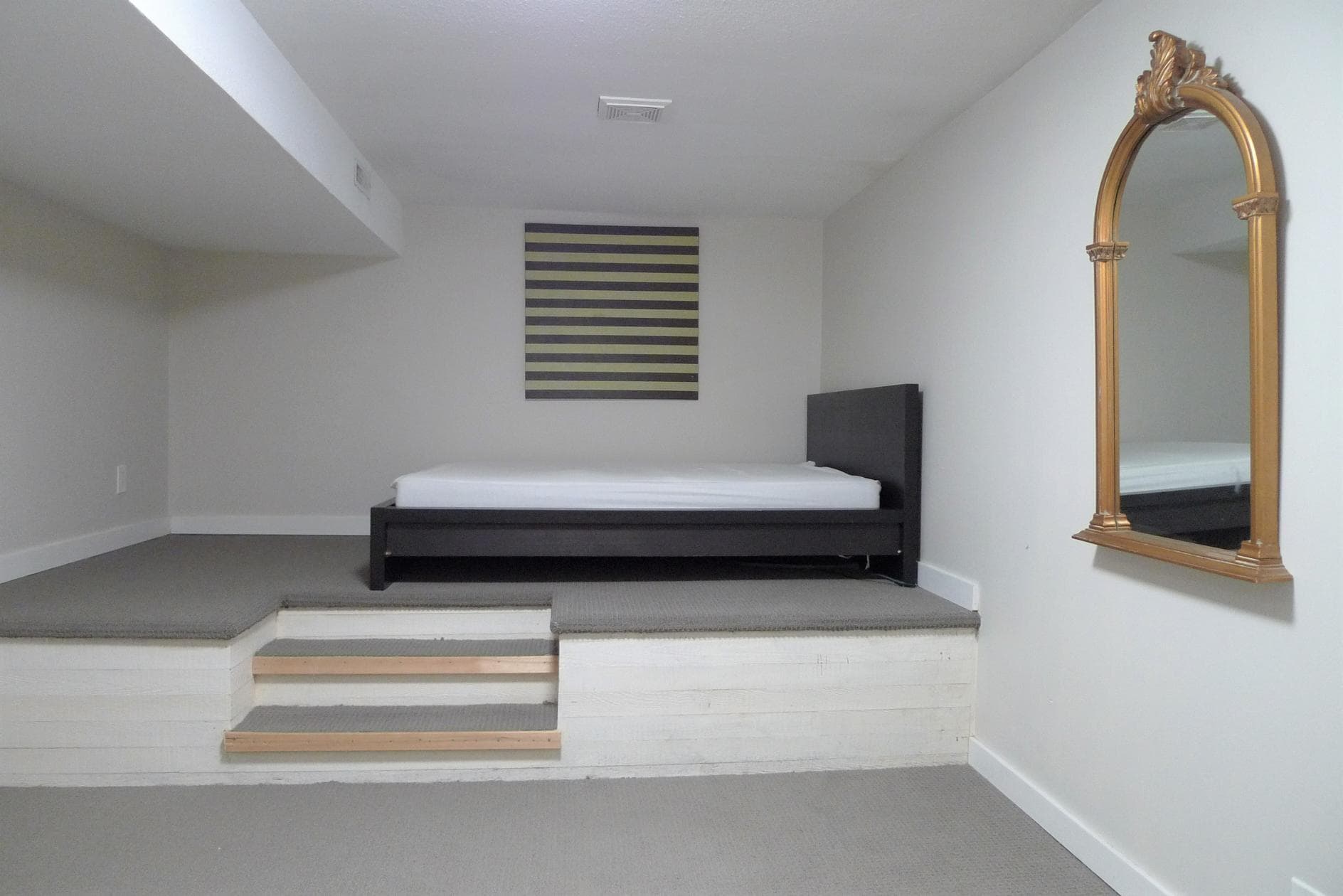 Spacious bedroom with a raised platform, modern black bed frame, elegant gold-framed mirror, neutral-toned walls, and contemporary artwork, ideal for Vancouver or BC real estate listings.