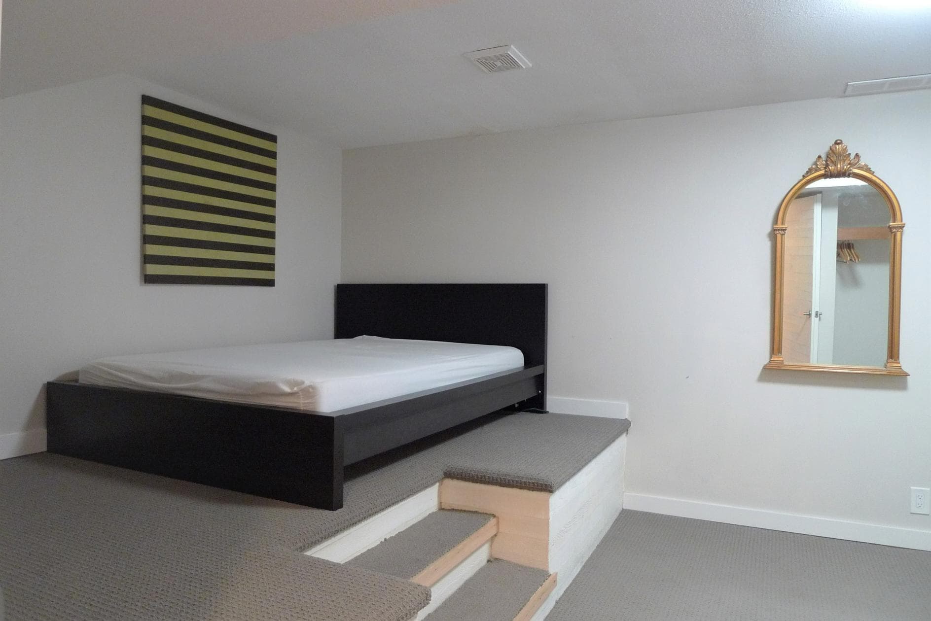 Affordable apartment bedroom with a raised platform, minimalist decor, black headboard, and decorative mirror, ideal for real estate listings by Orca Realty Inc.