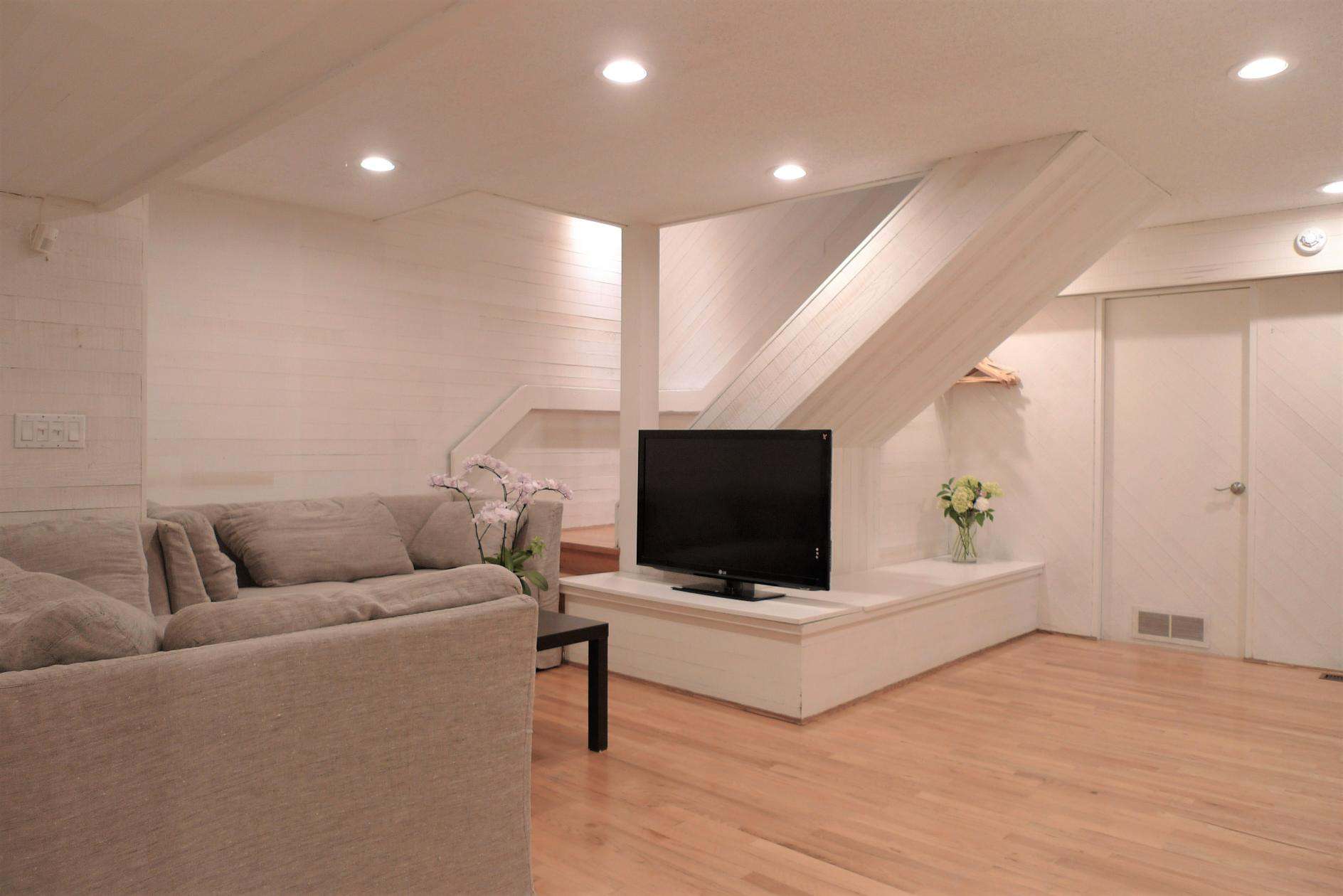 Modern living room with white wooden walls and flooring, cozy beige sofa, large flat-screen TV, decorative flowers, and recessed lighting, showcasing Orca Realty's rental and property management services in BC.