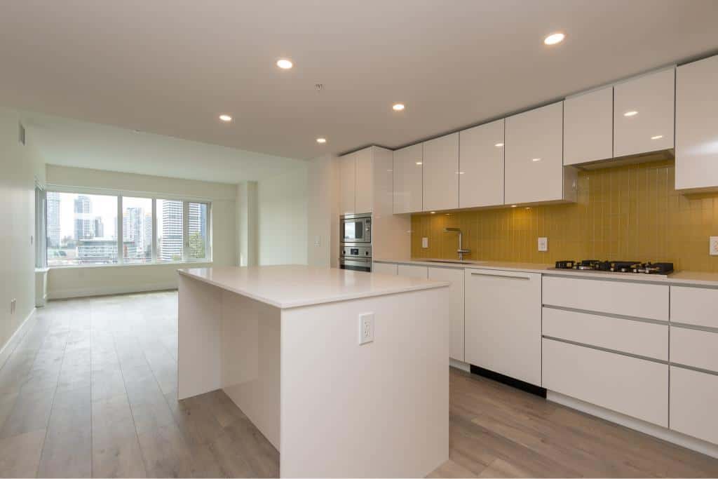 Bright modern kitchen with white cabinetry, stainless steel appliances, and a spacious open layout in a high-rise condo. Large windows offer city views and natural light. Unique yellow backsplash accents the contemporary design.