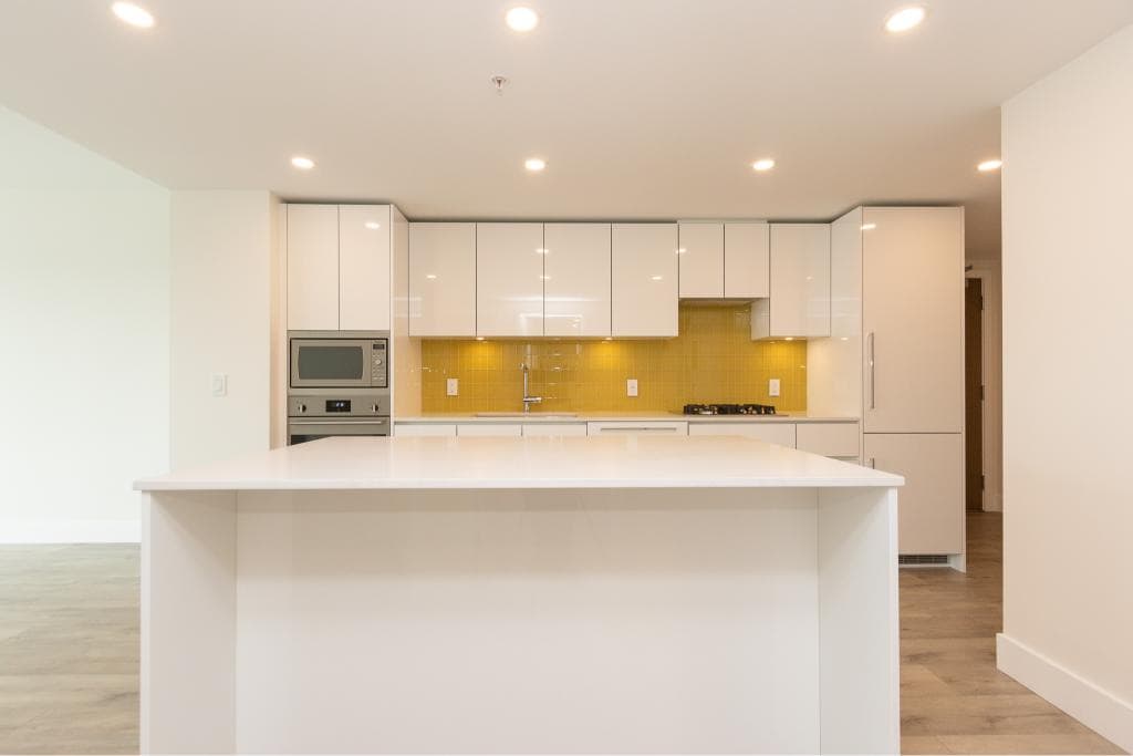 Modern white kitchen with yellow backsplash and stainless steel appliances, including oven and microwave, in a contemporary condo or home setting.