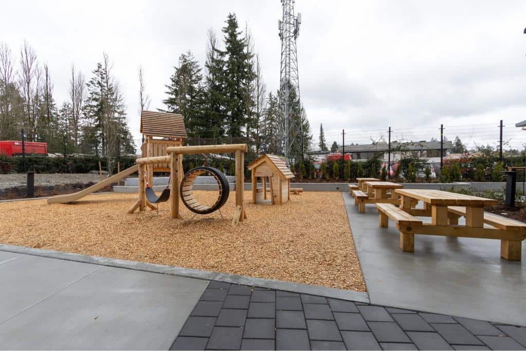 Natural wood playground equipment with slide, climbing circle, and small house, surrounded by outdoor seating and trees.