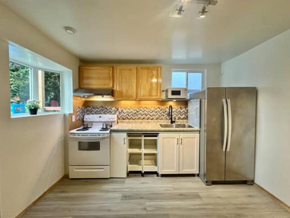 Modern small kitchen with wooden cabinets, stainless steel refrigerator, oven, microwave, granite countertop, and chevron backsplash in a bright Vancouver home.
