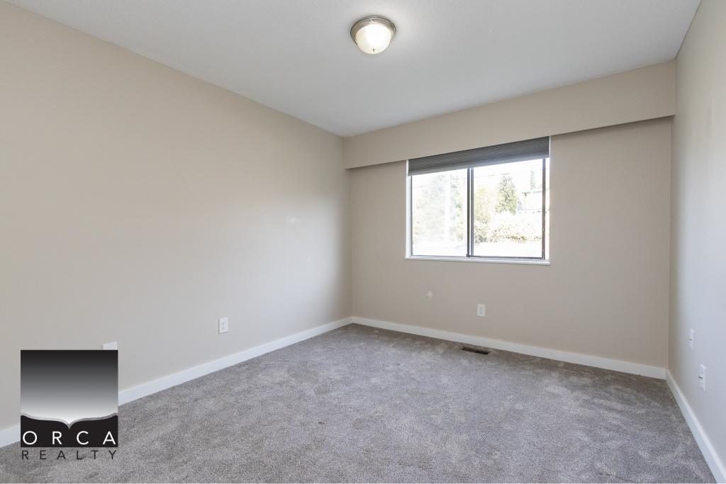 Bright empty bedroom with beige walls, grey carpet flooring, and large window in residential home - Orca Realty Inc.