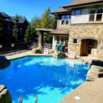 Sunlit outdoor heated swimming pool at Orca Realty Condominium, surrounded by lush trees and rustic stone building, perfect for relaxation and leisure.