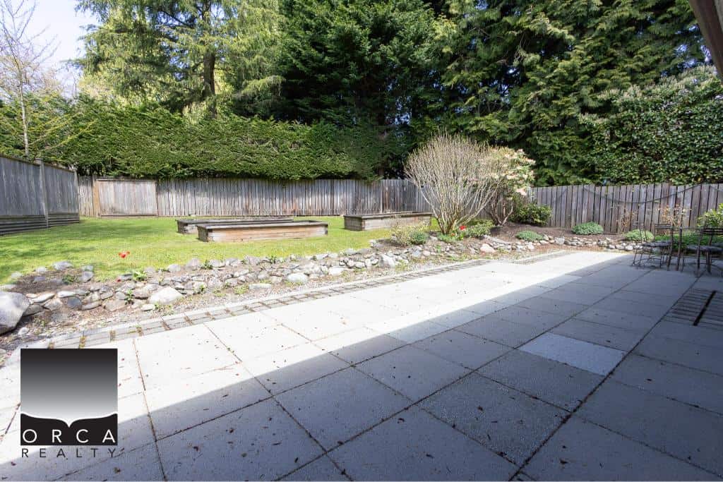 Lush backyard landscape with wooden deck, flowerbed, and mature trees, ideal for outdoor living and entertaining. Perfect outdoor space for Vancouver Island homes, featuring privacy fencing and natural garden design.
