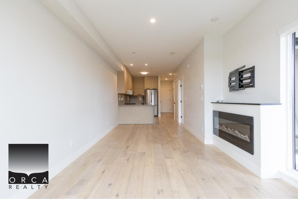 Spacious modern living room with light wood flooring, electric fireplace, and open kitchen in a new condo by Orca Realty Inc. in Vancouver, BC.