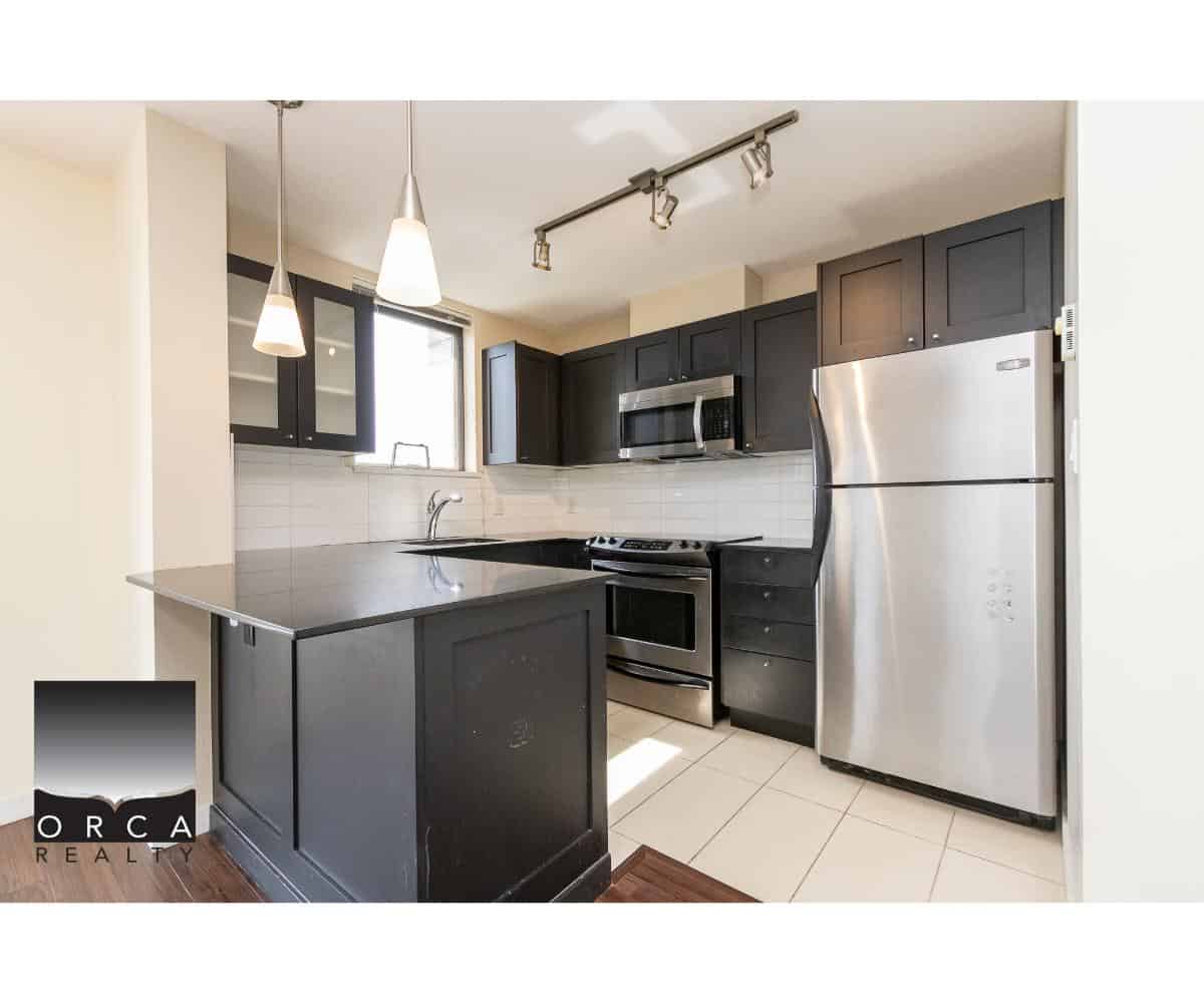 Contemporary black kitchen cabinets with stainless steel appliances, including a refrigerator, microwave, and oven, under modern pendant and track lighting. Perfect for Toronto-area homebuyers.