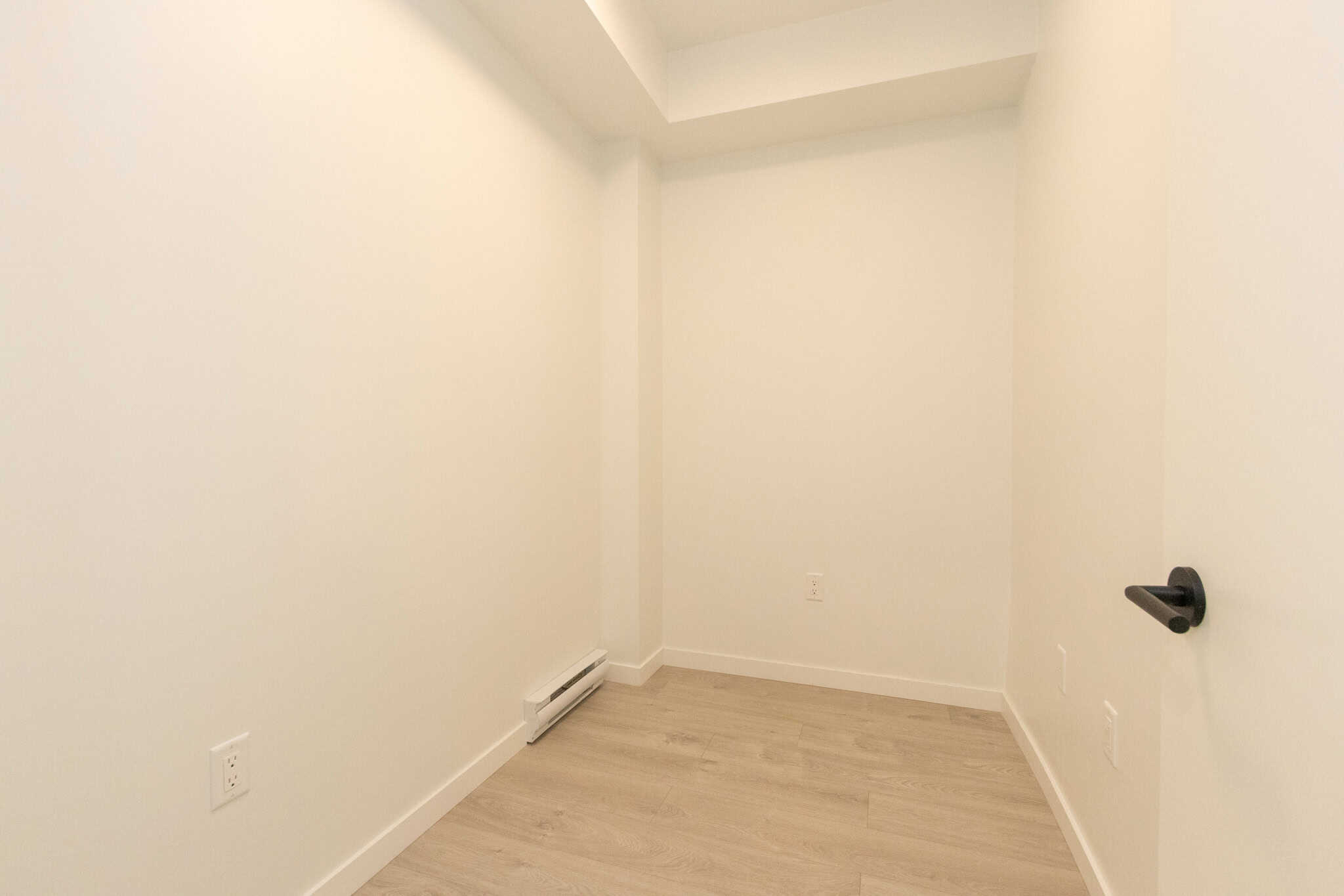 Bright, empty room with white walls and light wood flooring, ideal for a home office or bedroom, featuring a black door handle and baseboard heater, showcasing modern interior design.