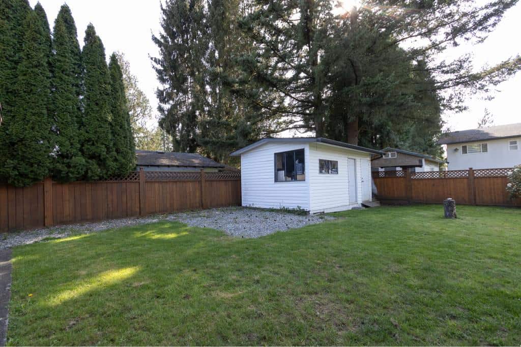 Fenced backyard with green lawn, mature trees, and a small white outbuilding, perfect for outdoor living and garden space, surrounded by tall trees providing privacy.