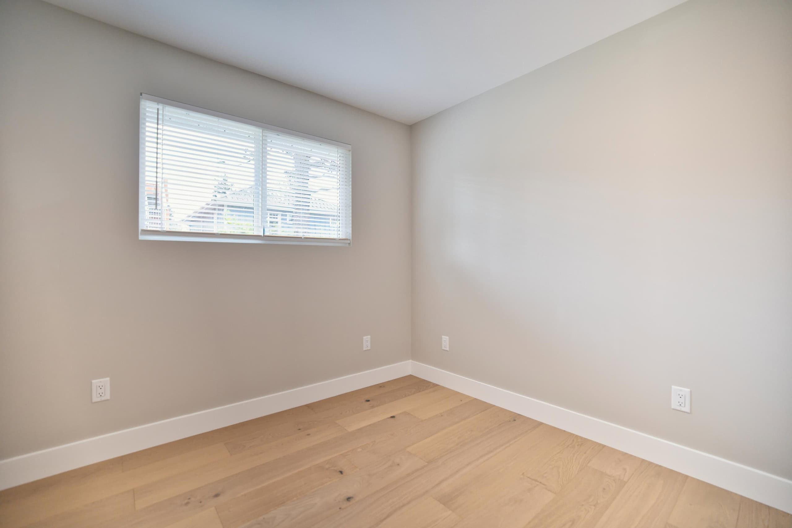Modern empty bedroom with large window, natural light, beige walls, white baseboards, hardwood flooring, ideal for Vancouver homebuyers, featured by Orca Realty Inc.