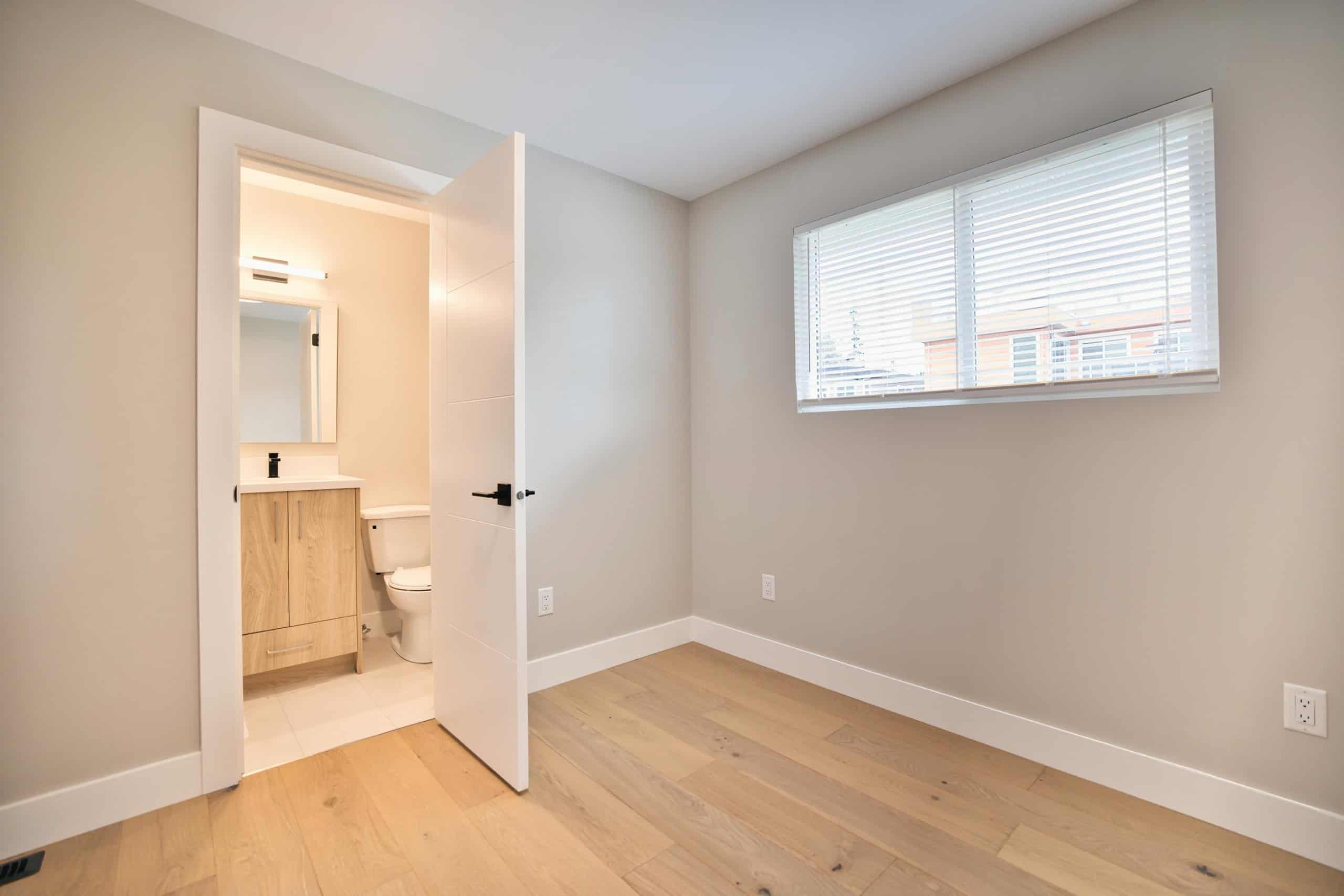Bright, empty bedroom with large window and attached bathroom, modern interior, light wood flooring, ideal for Vancouver or Victoria real estate listings.