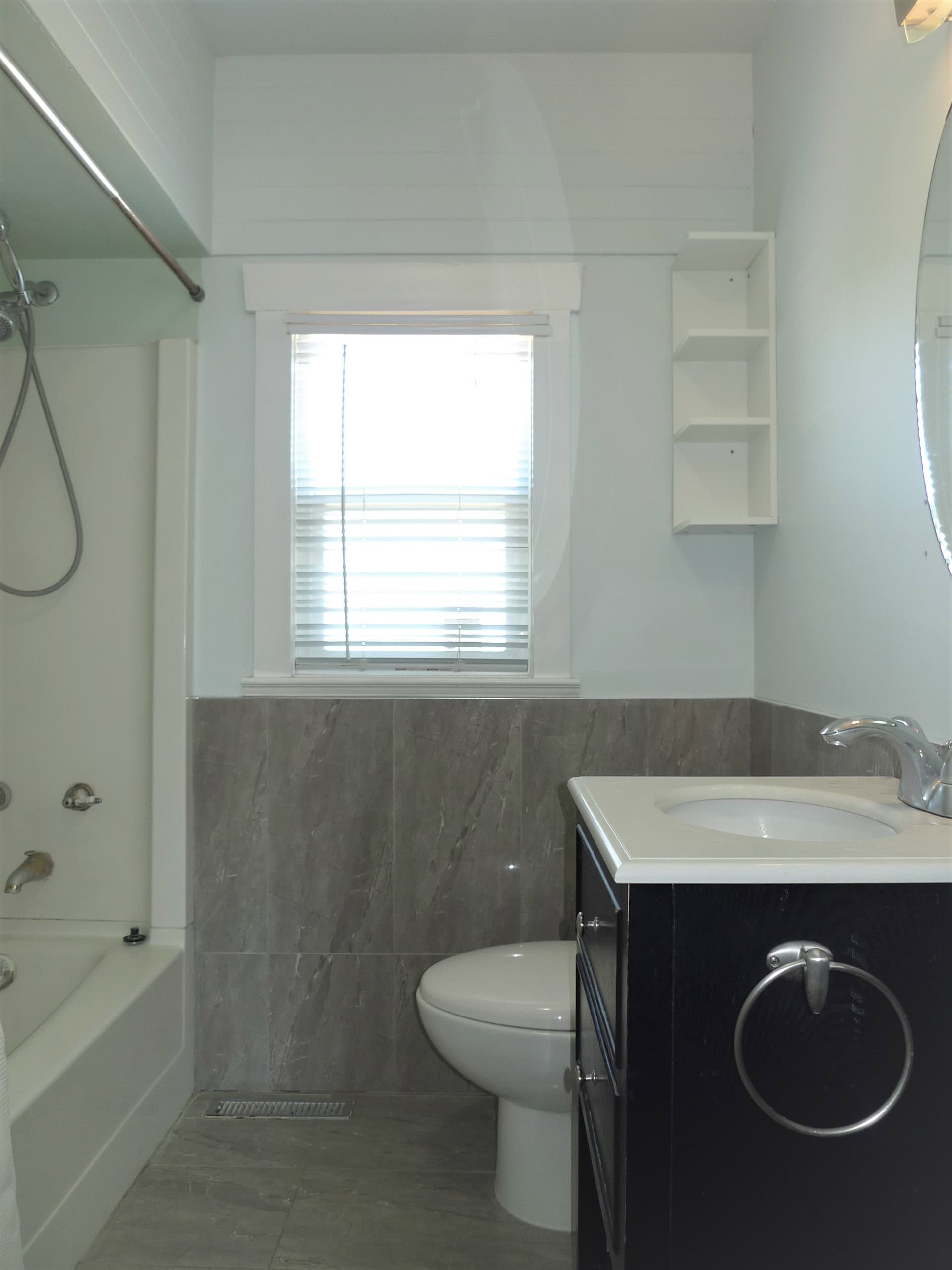 Bright modern bathroom with grey tile accents, white fixtures, and a window with blinds; featuring a walk-in shower, toilet, and black vanity for stylish, functional living space.