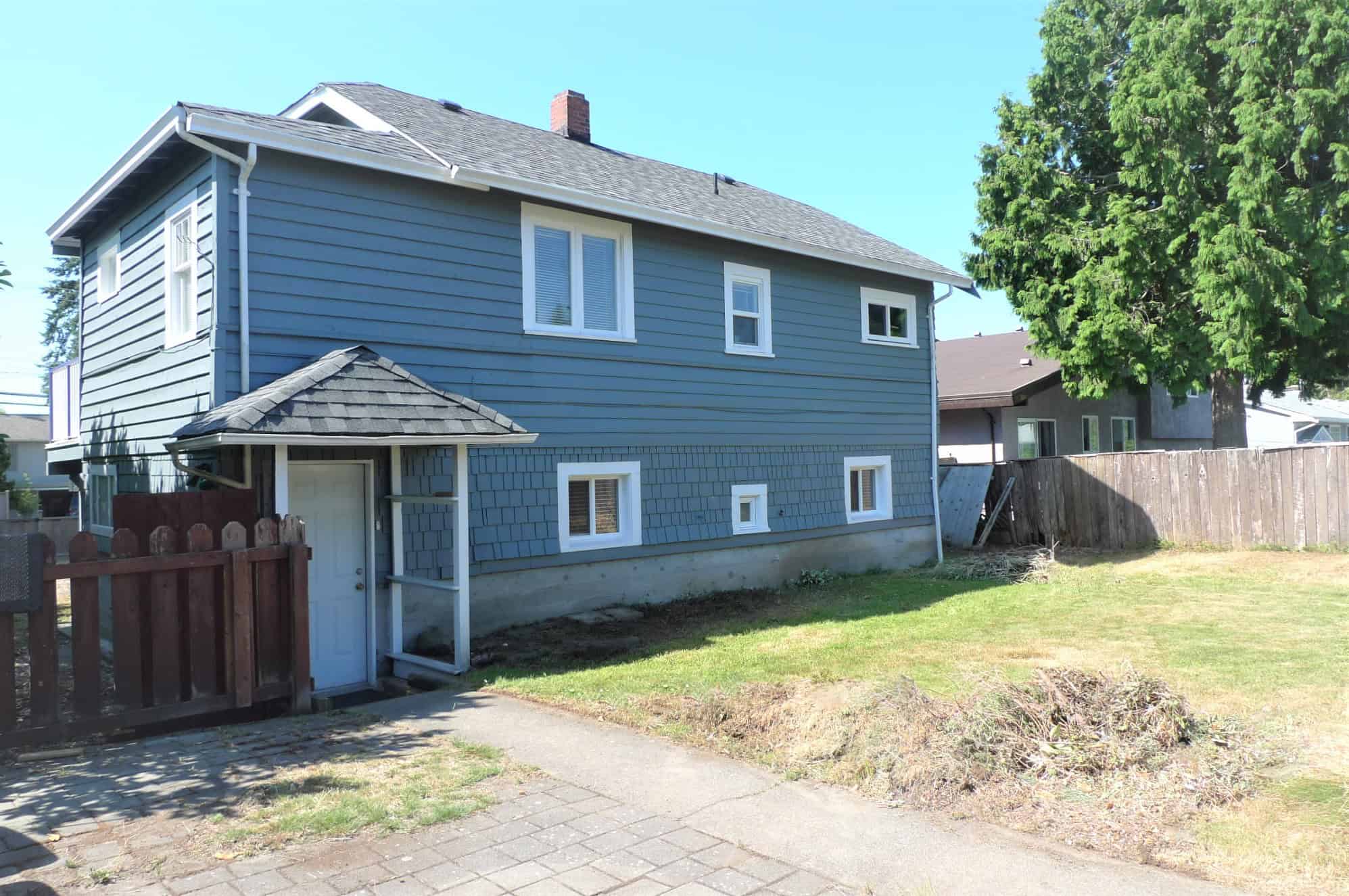 Blue two-story house with a well-maintained backyard in Vancouver, BC, with a wooden fence, mature trees, and a paved walkway, ideal for real estate investments and homes in the local market.