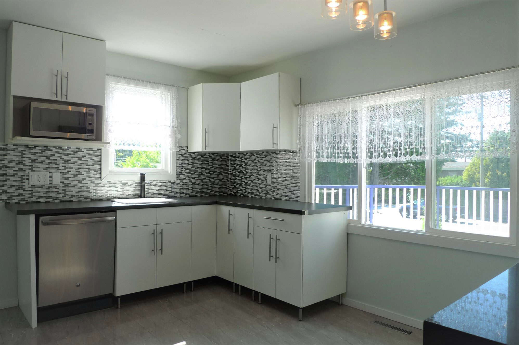 Modern kitchen with white cabinets, black countertops, stainless steel appliances, and a stylish mosaic tile backsplash, featuring natural light from large windows.