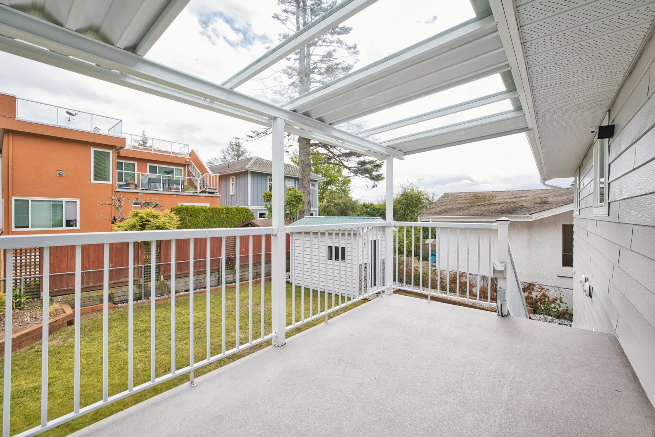 Spacious balcony with white railing and partial roof cover, overlooking a fenced backyard with lush grass, surrounded by family-friendly neighbourhood homes in Victoria, BC.
