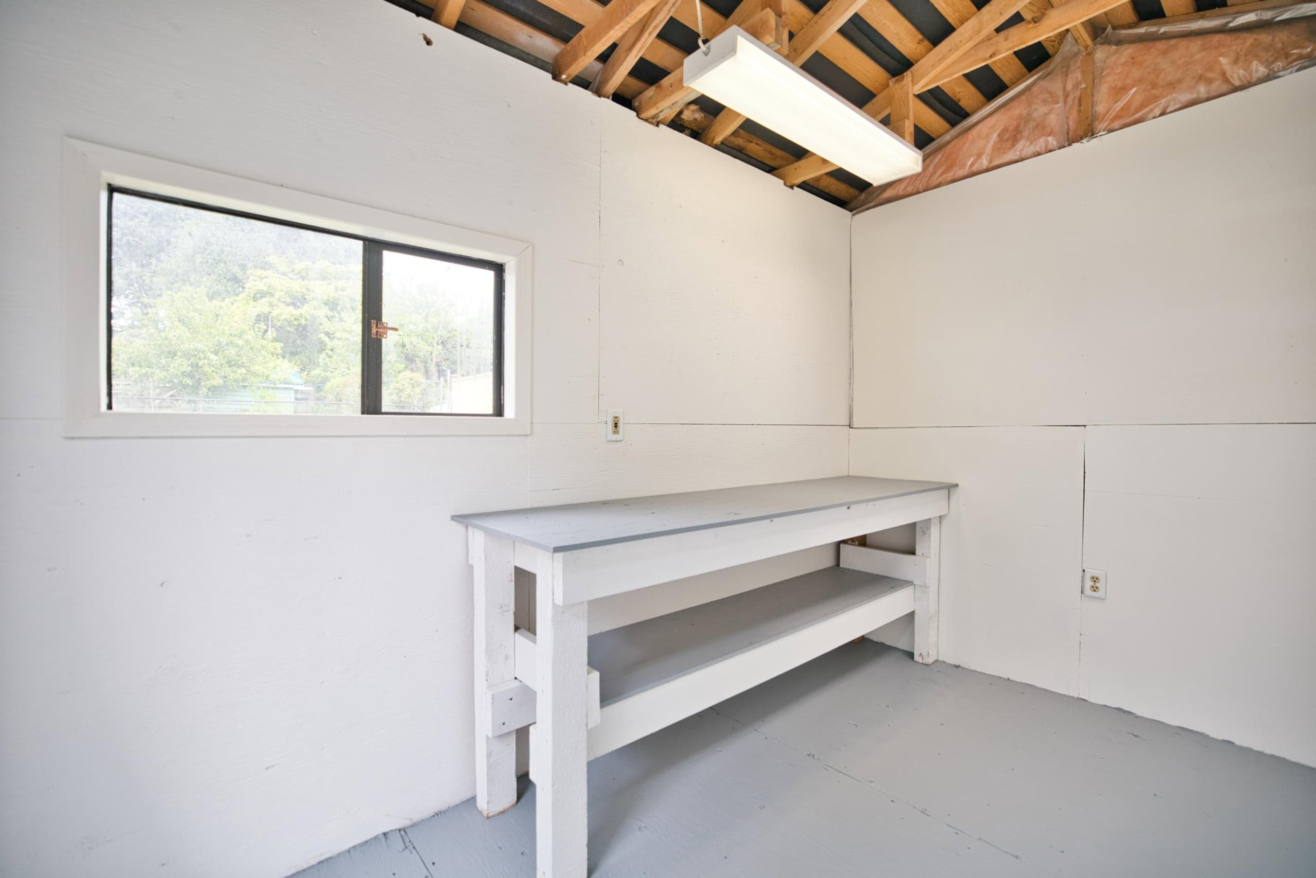 Bright unfinished workshop with white walls, a large window for natural light, and a sturdy workbench, ideal for home renovation projects, craft hobbies, or storage solutions in a residential property.