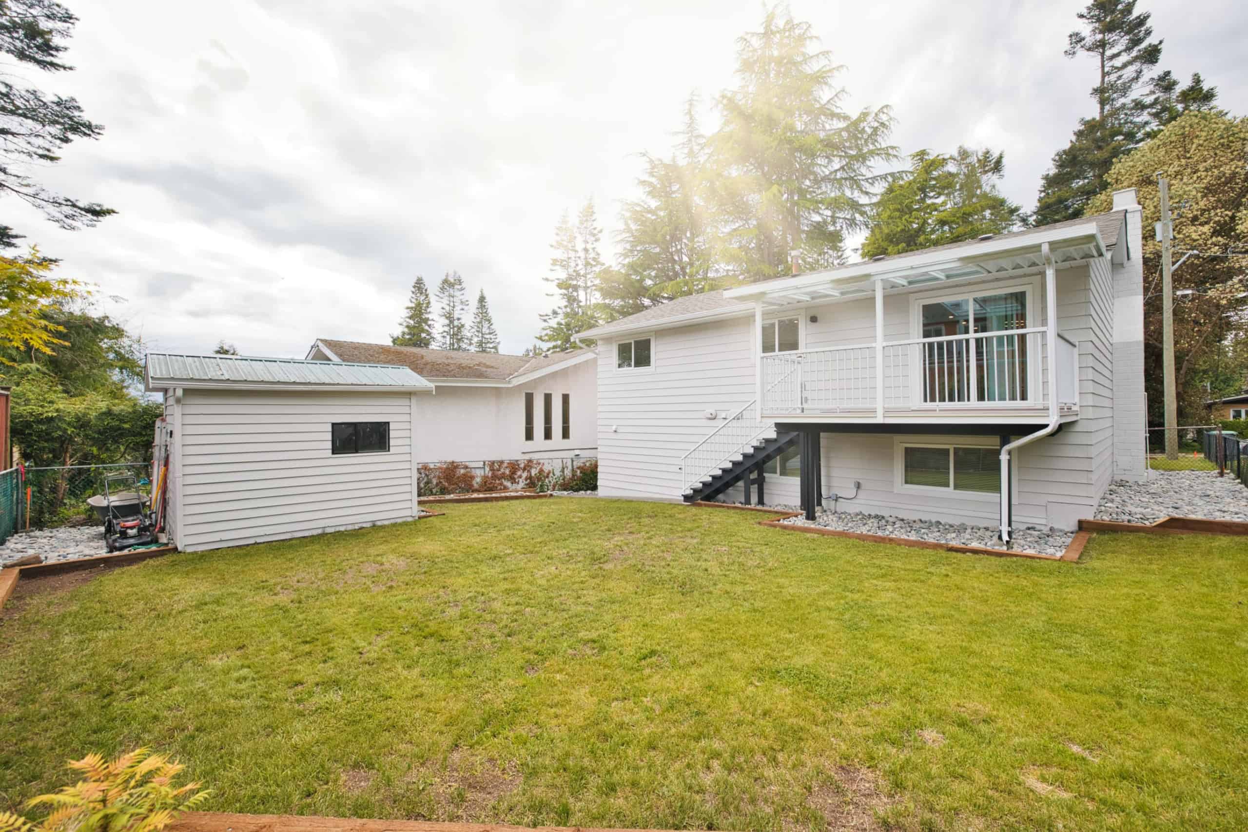 Bright white renovated backyard with a lawn, exterior stairs leading to a deck, surrounded by mature trees and built for outdoor living and privacy, ideal for buyers seeking a peaceful home.