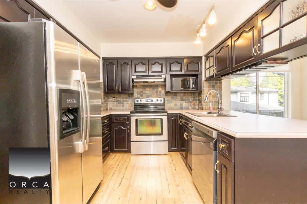 Stainless steel refrigerator, modern kitchen cabinets, and appliances in a bright contemporary kitchen with wooden flooring and backsplash.