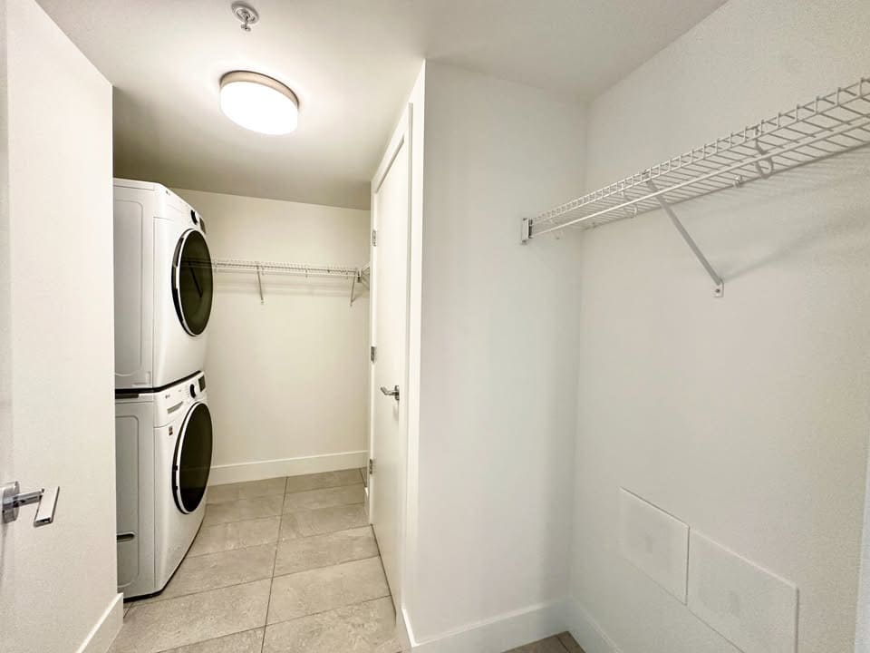 Modern laundry room with stacked washer and dryer, white wire shelving, neutral tile flooring, and clean white walls, perfect for convenient home living and organization.