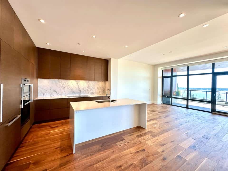 Spacious modern kitchen with wooden cabinetry, marble backsplash, and island in a high-rise apartment with floor-to-ceiling windows and stunning city views.