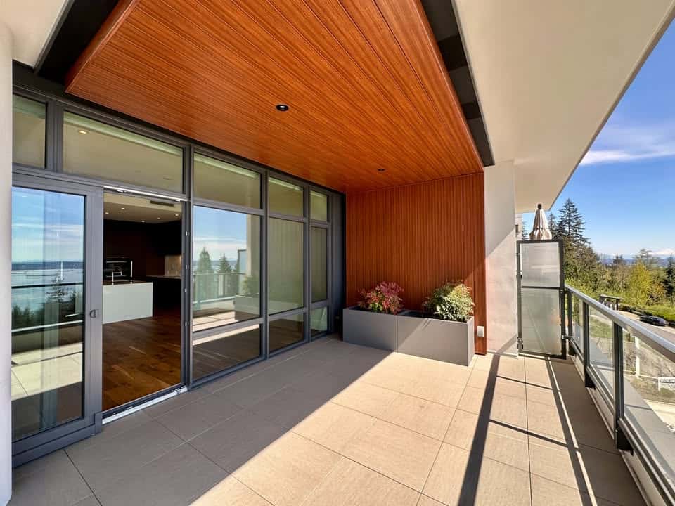 Balcony of modern residential property with glass sliding doors, wooden ceiling, and outdoor potted plants, overlooking lush trees and a scenic landscape.