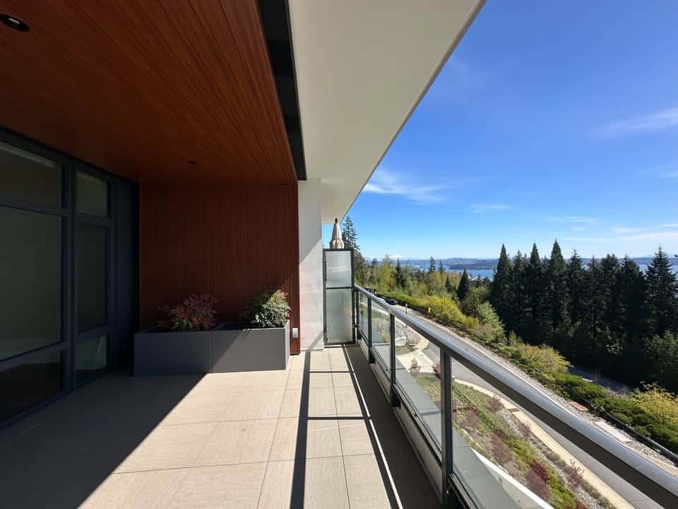 Spacious modern balcony with glass railing, overlooking lush green trees and a scenic water view, ideal for relaxing and outdoor entertaining in Vancouver.