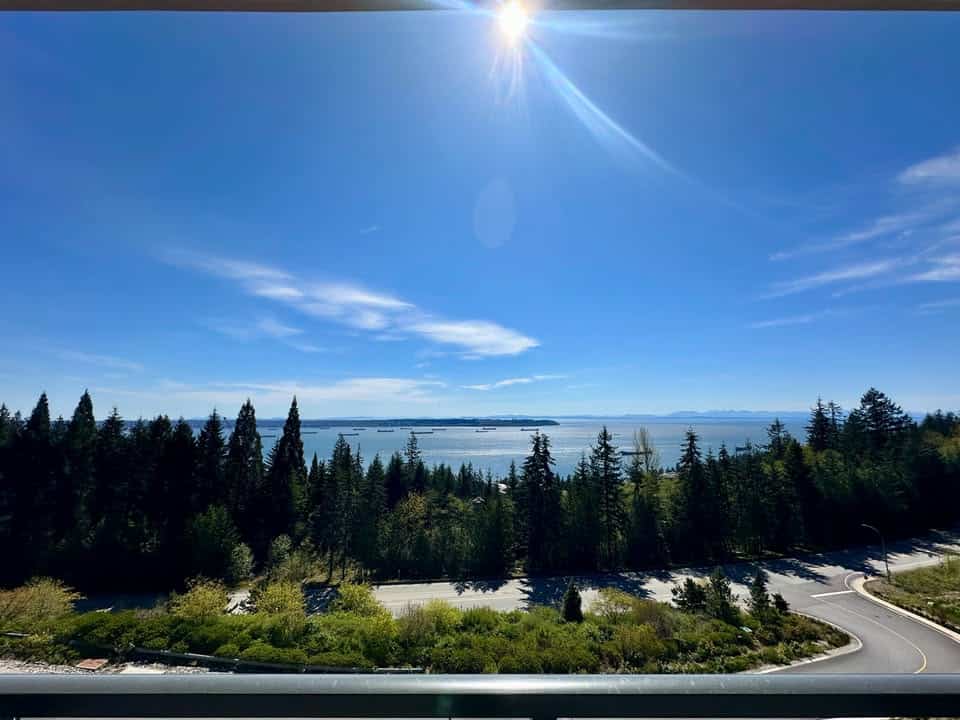 Breathtaking ocean view from a scenic location with lush green trees, a winding road, and a clear blue sky featuring the sun’s rays, ideal for real estate clients seeking waterfront or scenic properties in Vancouver.