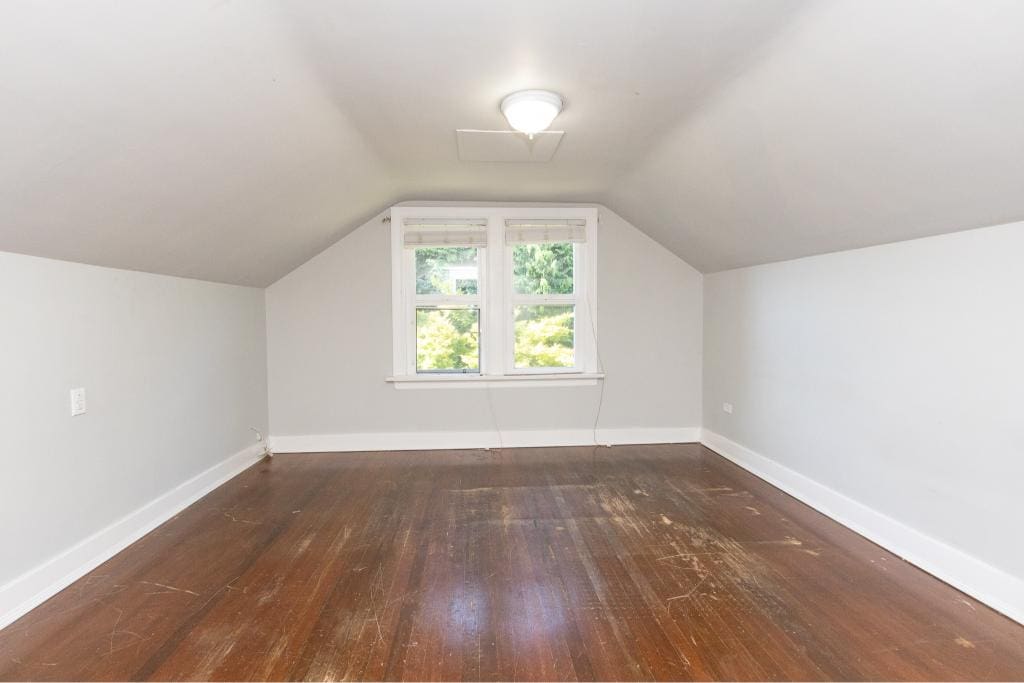 Bright empty attic bedroom with sloped ceiling, white walls, hardwood floors, and a window with lush green views, ideal for home staging or renovation projects - Orca Realty Inc.