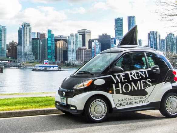 Smart black and white Eco-friendly electric car with "We Rent Homes" advertising parked by Vancouver waterfront skyline, representing Orca Realty Inc. real estate services.