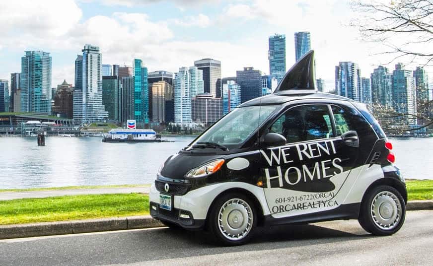 Smart black and white Eco-friendly electric car with "We Rent Homes" advertising parked by Vancouver waterfront skyline, representing Orca Realty Inc. real estate services.