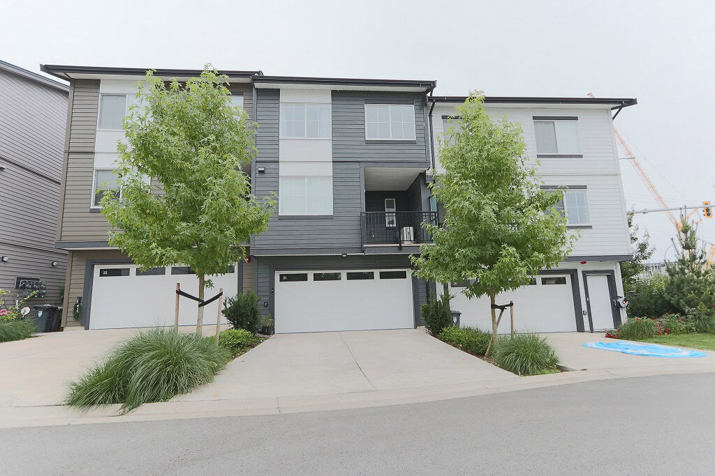 Modern multi-family townhouse building with grey and beige siding, adjacent to landscaped green spaces and trees, featuring secure garages and contemporary design in a residential neighbourhood.