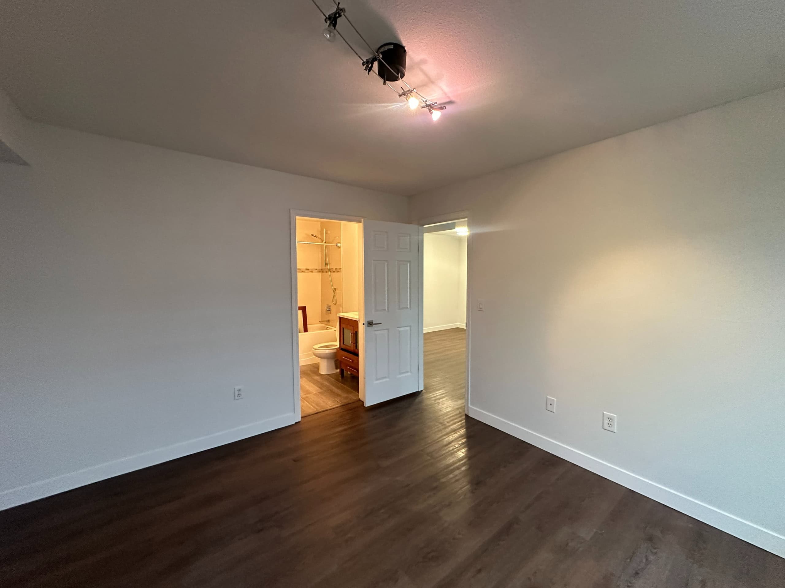 Modern empty bedroom with dark hardwood flooring, white walls, and a ceiling track light fixture, adjacent to a bathroom and hallway, ideal for open-concept living spaces.