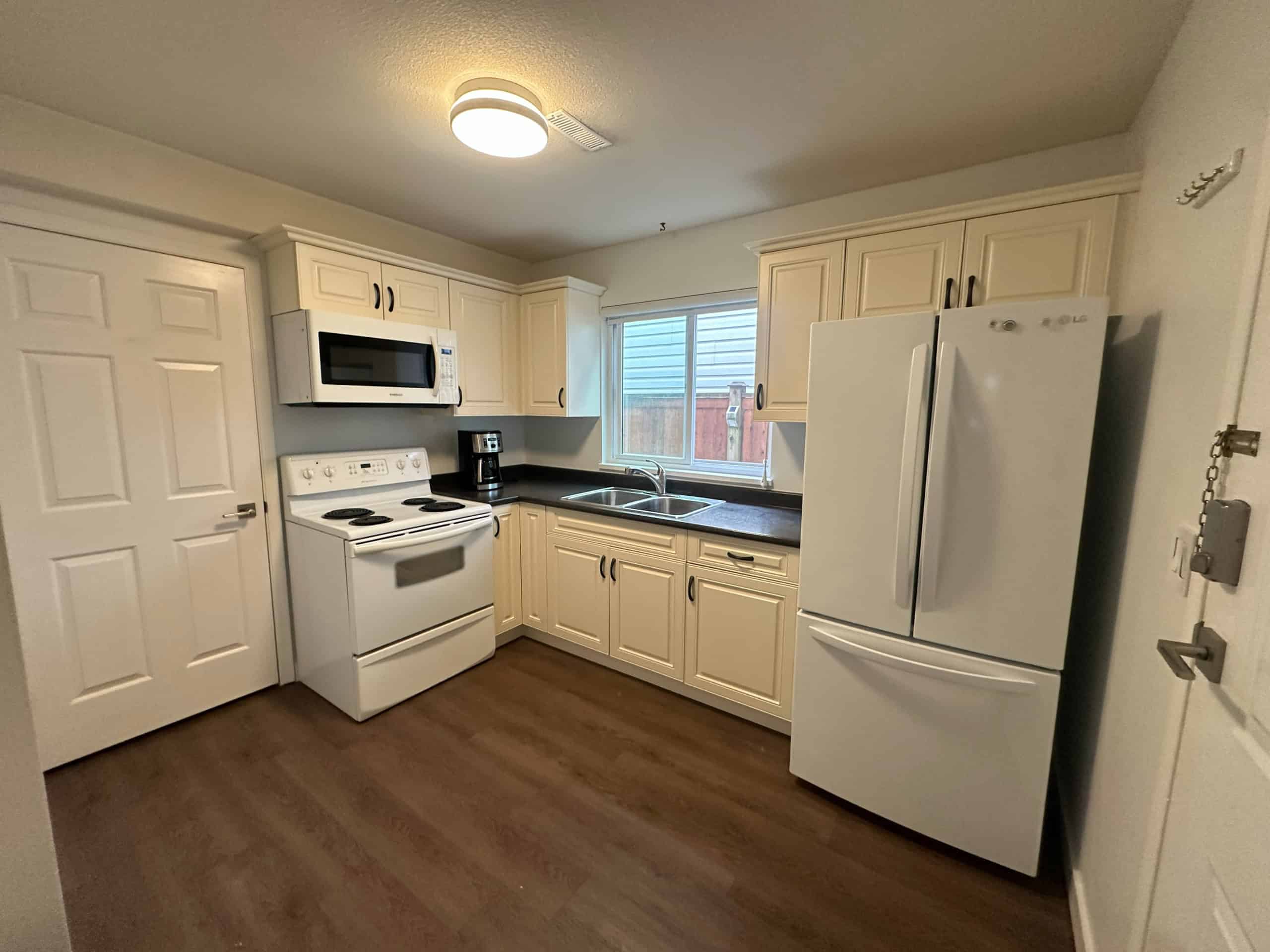 Bright modern kitchen with white cabinetry, black countertops, white appliances including fridge, stove, microwave, and coffee maker, stainless steel sink, and natural light from window, perfect for home living.