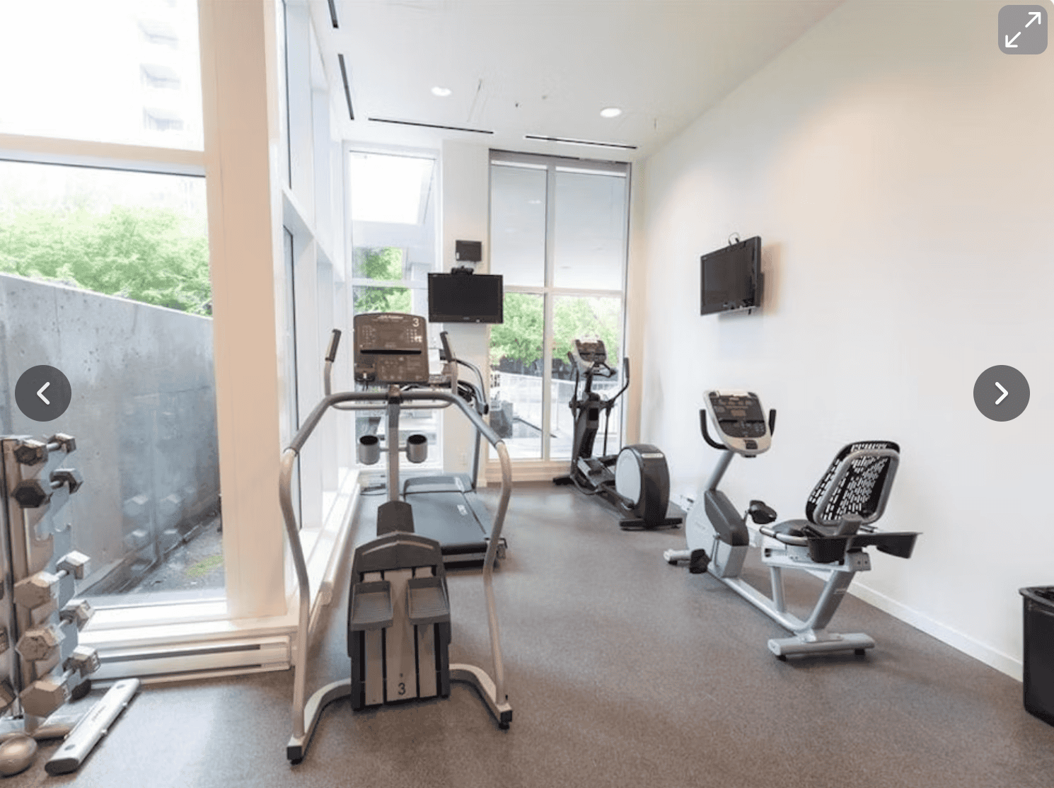 Modern fitness room with cardio equipment at Orca Realty Inc. featuring treadmills, stationary bikes, monitors, and large windows providing natural light.
