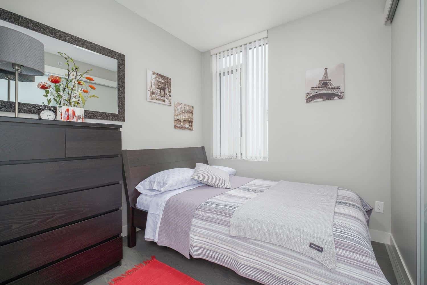 Cozy modern bedroom with minimalist decor, featuring a dark wooden dresser, a large mirror, and framed cityscape art, perfect for Vancouver real estate buyers seeking stylish homes in BC.