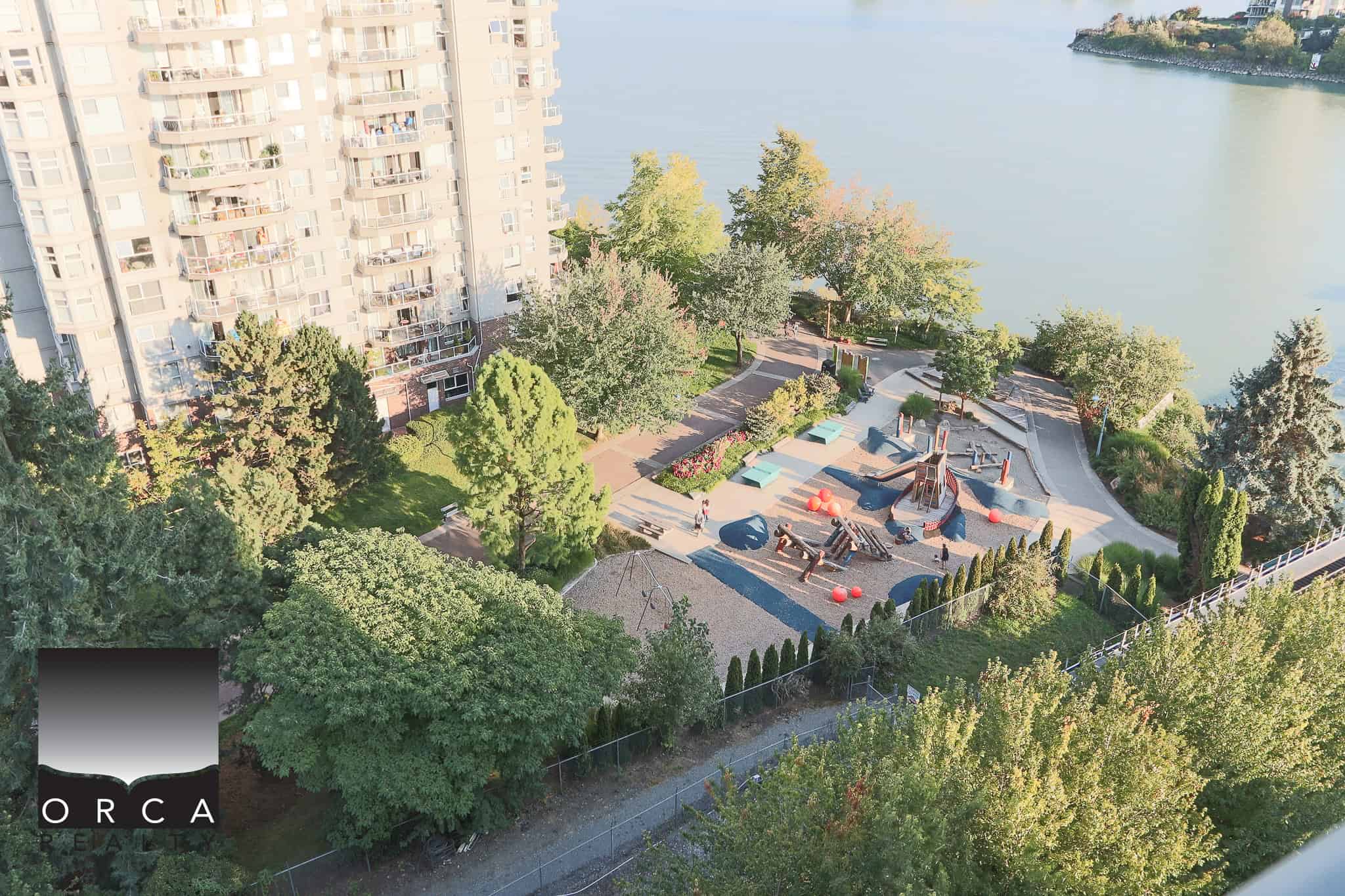 Modern waterfront condo building overlooking a lush park and playground area near the water in Vancouver, Canada.
