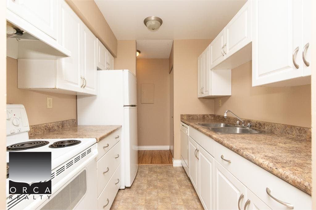 Modern kitchen with white cabinets, beige walls, and laminate flooring, featuring a refrigerator, oven, and double sink, ideal for Vancouver Island real estate listings.