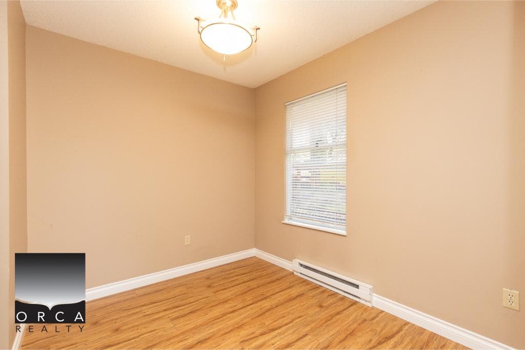 Bright empty bedroom with beige walls, hardwood flooring, and a window with blinds, showcasing a comfortable living space for Vancouver area real estate listings.