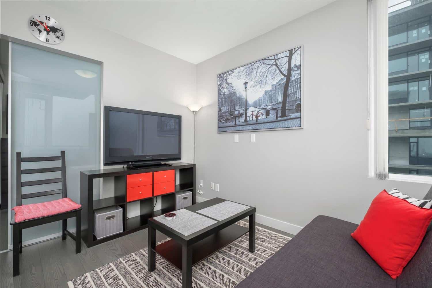 Modern living room with a flat-screen TV, stylish furniture, large window showcasing an urban view, perfect for condo or apartment rentals in Vancouver or Greater Vancouver.