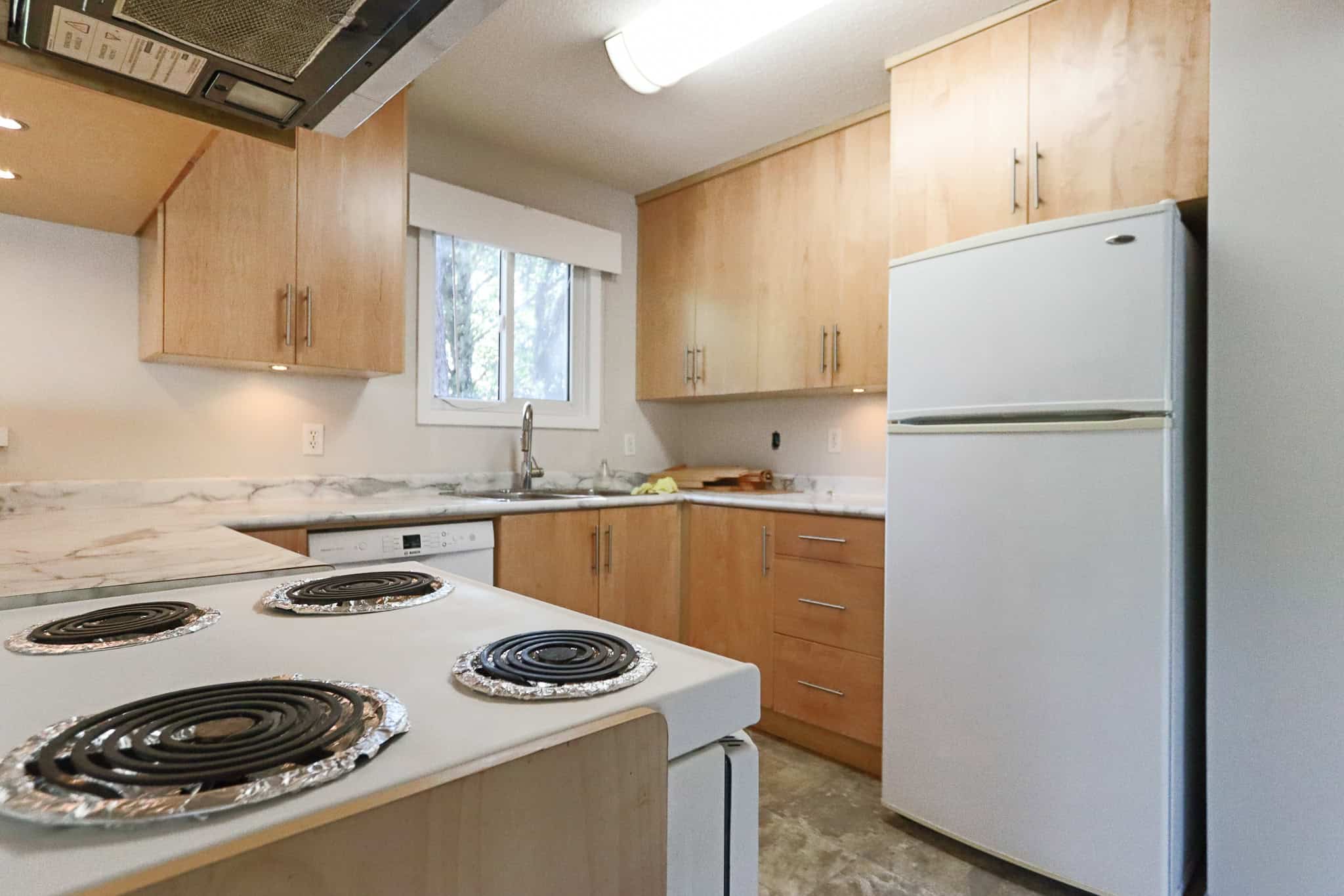 Modern kitchen with wooden cabinets, white appliances, and a marble countertop, featuring a stove, fridge, and a window with natural light, ideal for homebuyers seeking functional South Coast Vancouver Island homes.