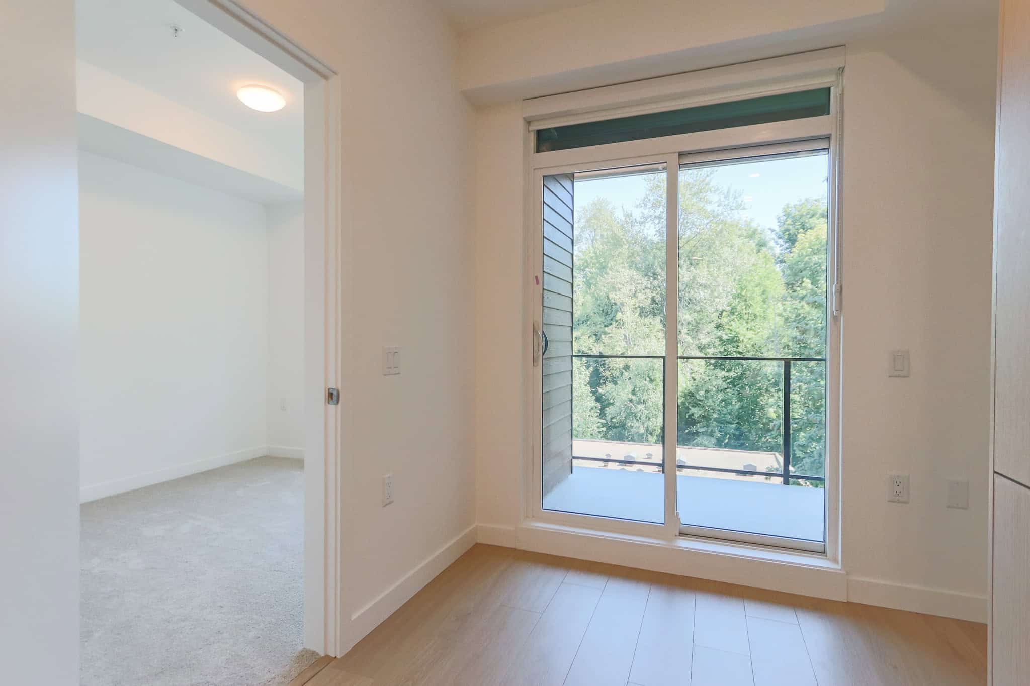 Bright interior featuring a large sliding glass door opening to a balcony with lush green trees, showcasing modern living spaces available through Orca Realty Inc.