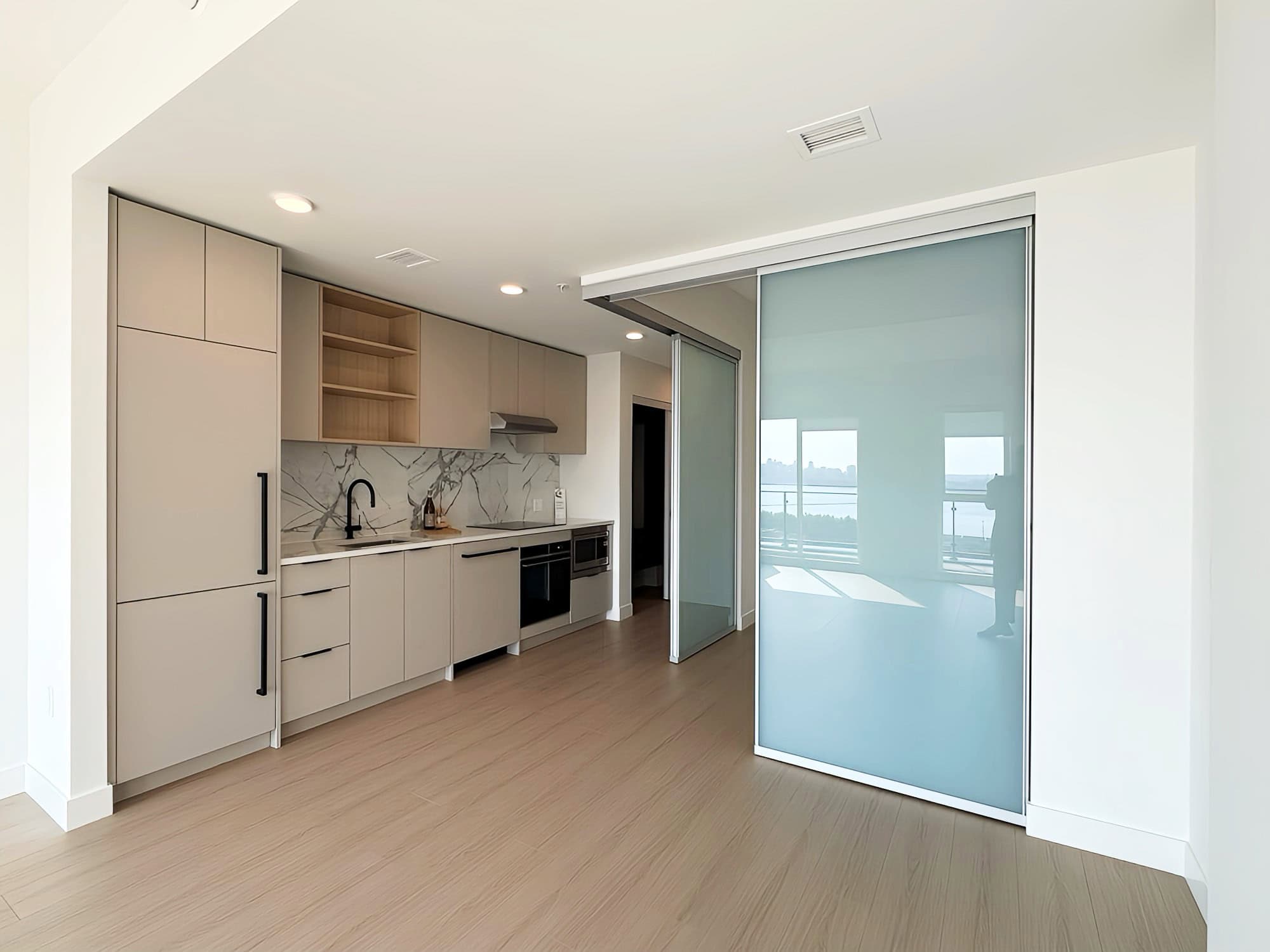 IMG_5598 Modern minimalist kitchen with beige cabinets, black hardware, marble backsplash, and sliding glass door leading to balcony overlooking scenic waterfront views.
