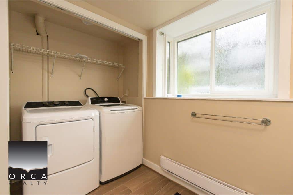 Clean laundry room with washer and dryer, bright natural light from large window, beige walls, wall-mounted towel bar, and organized shelving, ideal for Vancouver Island property.